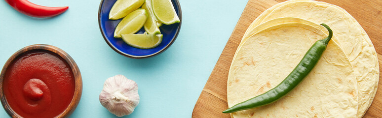 Canvas Print - Top view of tortillas, tomato sauce with garlic and lime slices on blue background, panoramic shot