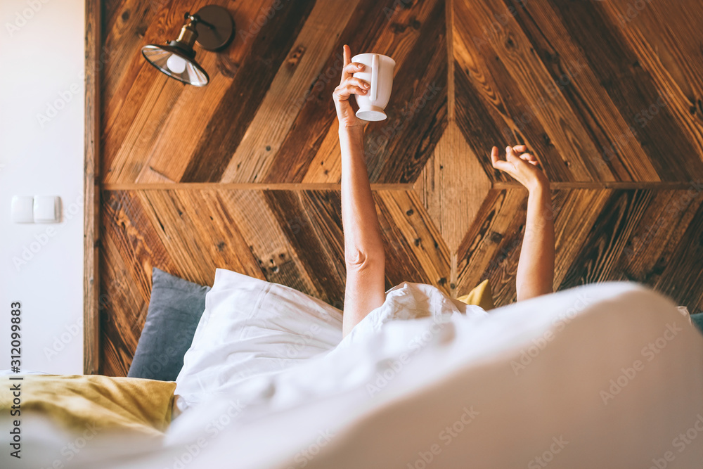 Lazy female lying under the white blanket on the  linen bed and holding the fresh coffee cup in the early morning. Lazy day off and "coffee in bed"concept image. - obrazy, fototapety, plakaty 
