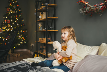 cute little girl sitting on the bed with a teddy bear near a christmas tree.