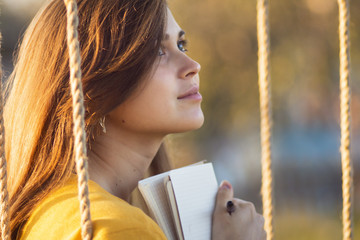 Wall Mural - cute girl on a swing with a notebook and pen keeps a diary of feelings in nature, a woman composes and writes, romantic mood