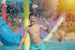European boy splashing in pool at waterpark against colorful plastic slides. He smiling and looking into camera.