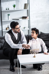 Wall Mural - smiling jewish father and son holding candle in apartment