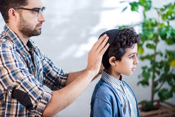 Wall Mural - side view of jewish father wearing hat on son in apartment