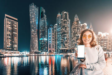 Wall Mural - Happy asian girl with smartphone in her hand walking and exploring the grand Marina district in Dubai with the Bay and skyscrapers at night