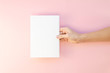 Women hand hold white template paper or magazine on pink background. Mockup flat lay with copy space