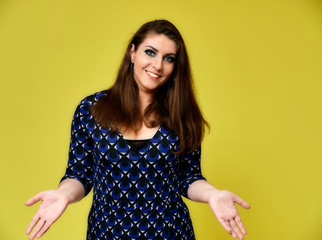 Portrait of a pretty brunette woman with a beautiful hairstyle and with excellent makeup in a dark blue blouse on a yellow background. It stands showing hands with a smile in front of the camera.