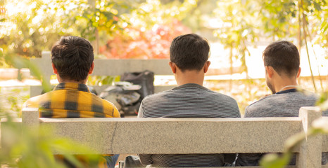 Back view of three friends sitting on table busy on mobile at park - Concept of best friends and togetherness.