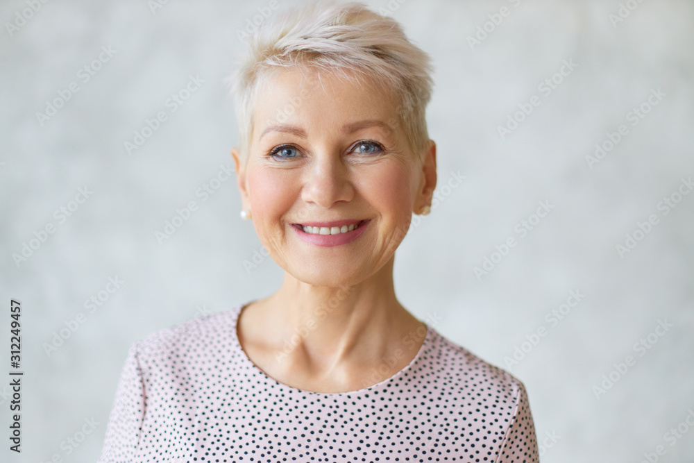 Close up image of good looking beautiful mature blonde female with blue eyes, elegant make upand pixie hairstyle smiling at camera, having confident happy facial expression, being in good mood - obrazy, fototapety, plakaty 