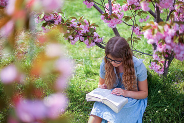 Pretty young school girl in glasses reading book near blossom sakura tree in garden. Adorable teenager learning in spring park