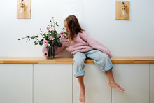 Little Barefoot Girl In A Pink Oversized Knitted Sweater Sitting On A Shelf In Corridor At Home, Hugging Glass Jar With Roses In It. Her Feet Hanging In The Air. Sleeves Are Far Too Long For Her.
