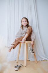 Wall Mural - Cheerful little girl in a beige knitted sweater sitting on a stool at home, in front of a curtain. She's taking leopard sandals off of her bare feet. Top view.