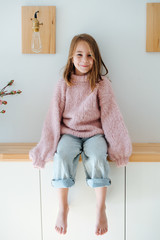 Wall Mural - Happy smiling cute little barefoot girl in a pink oversized knitted sweater sitting on a shelf in corridor at home. Her feet hanging in the air. Sleeves are far too long for her.