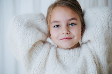 Wall Mural - Portrait of a brunette little girl in white fluffy knitted sweater putting her hair in a pony tail. At home, in front of a curtain. She's looking at camera. Close up.