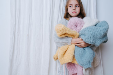 Wall Mural - Sad unhappy little girl is hugging pile of soft fluffy multi-colored freshly washed sweaters, similar to the one she's wearing. At home, in front of a curtain. Doing chores. half length.