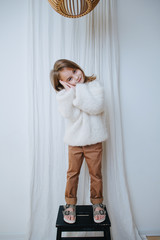 Wall Mural - Playful little girl in white fluffy knitted sweater is standing on a stool, making sleep gesture. At home, in front of a curtain. Full length.