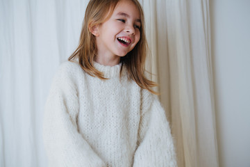 Wall Mural - Portrait of a cheerful laughing brunette little girl in white fluffy knitted sweater. At home, in front of a curtain. Half length.
