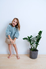 Wall Mural - Laughing little barefoot girl in a blue sweater standing on a parquet next to the plant near room wall. At home. full length.
