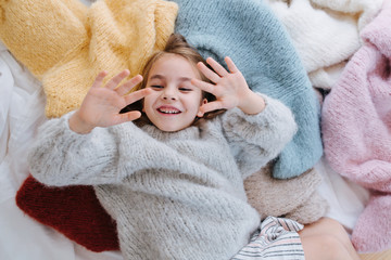 Wall Mural - Happy giggling littlegirl is lying on a pile of soft fluffy multi-colored freshly washed sweaters, similar in fashion to the one she's wearing. Playing with some one, defending with hands. Top view.