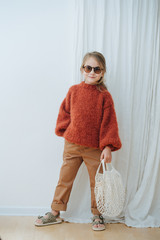 Wall Mural - Cute smiling little hipster girl in sunglasses, dark orange knitted sweater, brown jeans, leopard sandals holding net bag. With hand in a poket. Full length.