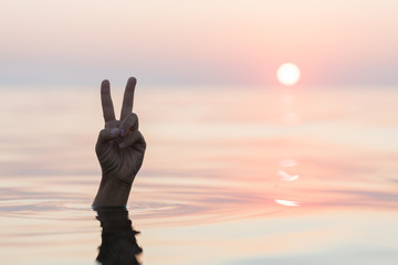 Hand emerging from the sea showing a peace sign in sunset