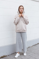 Wall Mural - Beautiful young girl in fashionable biege hoodie and pants on light background. Walking on the street.