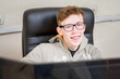 A cheerful guy with glasses and braces on his teeth works at a computer. A smiling teenager is sitting in an office chair behind the monitors. Portrait of a student with red hair.