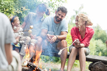 Happy Family Sitting With Woman While Roasting Marshmallows Over Burning Campfire At Park