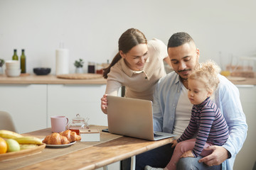 Warm-toned portrait of modern mixed race-family using computer while sitting in cozy kitchen interior with cute little daughter, copy space