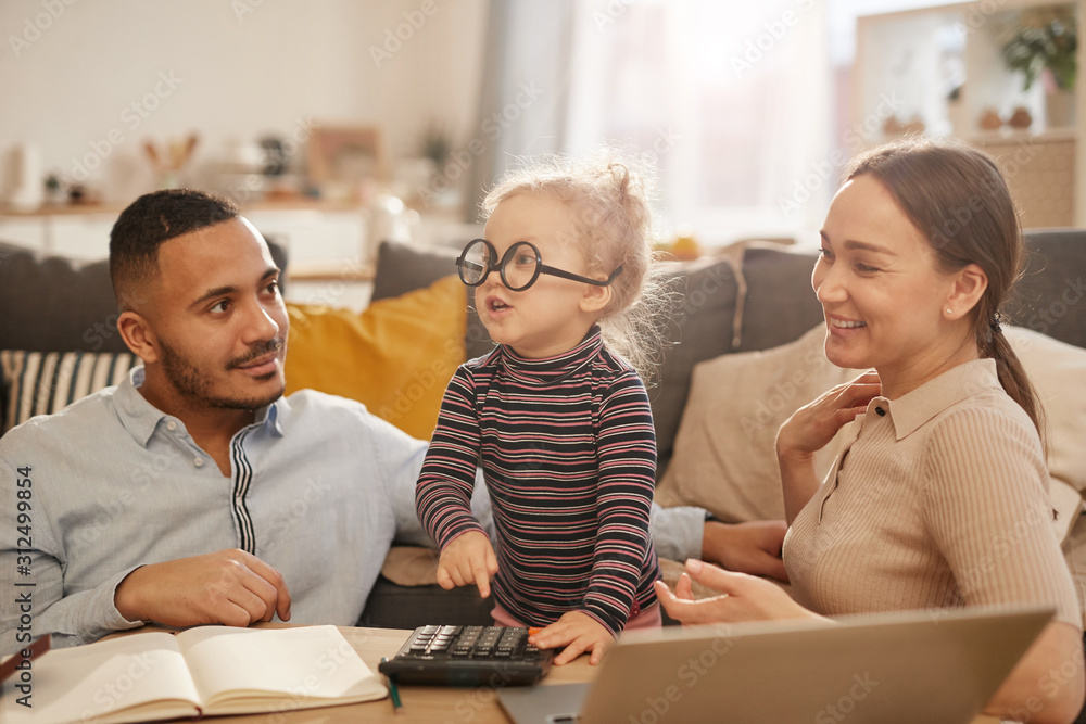 Portrait of happy young family counting home finances with cute little girl wearing glasses in sunlit apartment - obrazy, fototapety, plakaty 