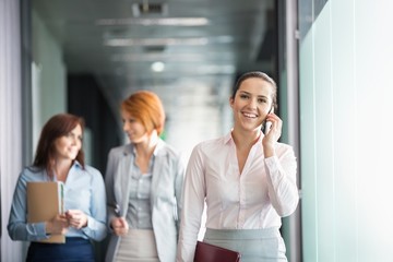 Wall Mural - Young businesswoman on call with colleagues in background at office corridor