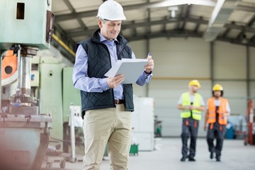 Wall Mural - Mature male inspector writing on clipboard while workers in background at factory