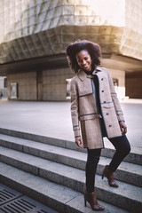 Beautiful african american young woman with afro and large hoop earrings in a stylish coat, smiling. Urban street portrait.