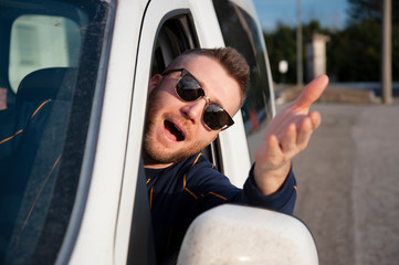 Young man with sunglasses comes out of the car window with his head with his arm raised. Face expression of angry driver arguing and gesturing. Express courier with work vehicle. Negative emotions.