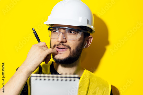 Fotomural Studio portrait of a young thinking man, builder engineer wearing safety helmet and glasses for construction on yellow background