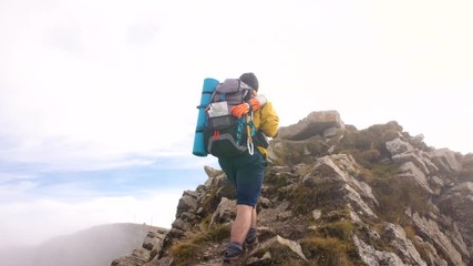 Wall Mural - young man hiker on mountain with cloudy sky and fog. yellow jacket, backpack and beanie. traveling d
