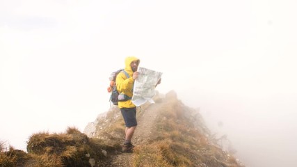 Wall Mural - young man hiker on mountain trail reading map. cloudy sky and fog. yellow hood jacket and backpack. 