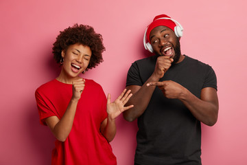 Wall Mural - Amused curly haired woman pretends singing in microphone, dances carefree together with boyfriend, enjoy music, dressed casually, isolated over rosy wall. People, fun, entertainment concept.