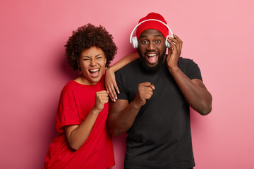 Sticker - Youth lifestyle and party concept. Joyful dark skinned woman and man sing songs loudly, have overjoyed expressions, dance together, use headphones, isolated over rosy background. Music, technology