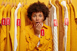 Emotional stupefied young woman with Afro hairstyle covers mouth, shocked by big sales and discounts, selects shirt from rack at clothing store, stands in stupor. Row of clothes with red tags
