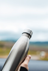 Close-up of female hand holding a steel reusable thermal water bottle on background of sky.