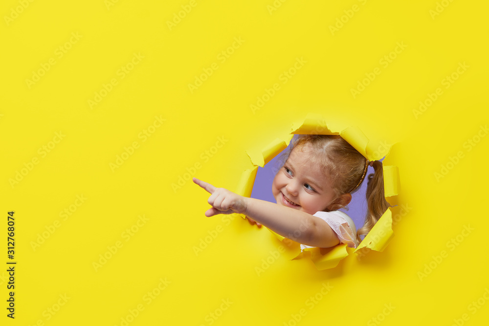 Little surprised child looking, peeping through the bright yellow paper hole. Showing hand to side. Advertise childrens goods. Copy space for text. - obrazy, fototapety, plakaty 