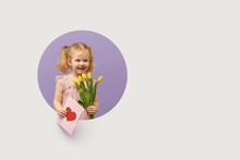 Adorable Smiling Child With Spring Flower Bouquet And Greating Card In Circle Hole In The Wall. Little Toddler Girl Holding Yellow Tulips As Gift For Mother. 8 March Or Mother Day Concept