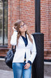 © Марина Демешко - A cute girl in the white jacket with glasses  is adjusting her hair and looking sideways with shy smile while standing outdoors on the background of a brick wall. Dressed in business style