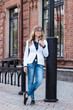 © Марина Демешко - A pretty girl in a white jacket and blue jeans with glasses looking sideways and standing outdoors on the background of a brick wall. Dressed in business style. Vertical portrait