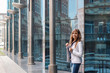 © Марина Демешко - A european young businesswoman in a white jacket is smilng, is standing against the background of a mirrored blue business building, the prospect of the pillars. Dressed in business style