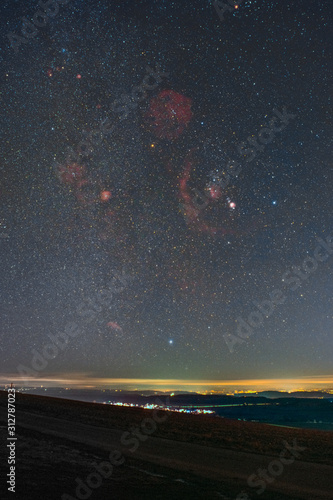 Fotomural Winter night sky including Barnard’s Loop, the Orion Nebula, the Flame Nebula, the Rosette Nebula, and the bright star Sirius photographed from the summit of the mountain Witthoh in Germany
