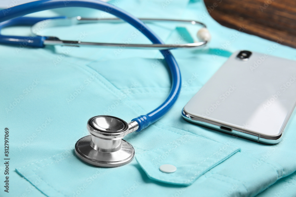 Stethoscope, mobile phone and doctor's uniform on table, closeup