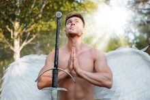 A Warrior Angel, A Bodybuilder With Wings Behind His Back, Stands In A Ray Of Light Next To A Two-handed Sword With His Hands Folded In Prayer.