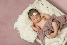 Sleeping Newborn Baby Girl Laying On A Cream Colored Blanket On A Pink Purple Backdrop With A Headband In Her Hair