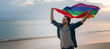 © olezzo - Cheerful guy with a rainbow flag on the beach. Young man holding a rainbow flag against the ocean sky